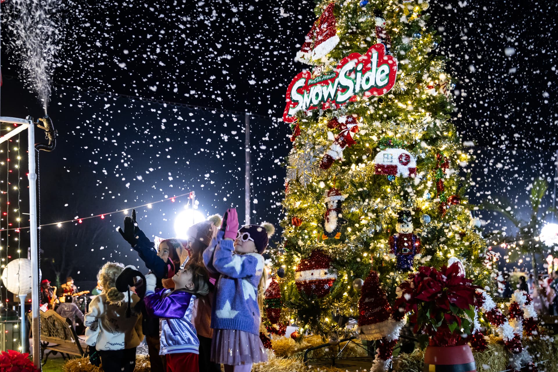 Kids reaching for falling snow beside the giant lit SnowSide Christmas tree